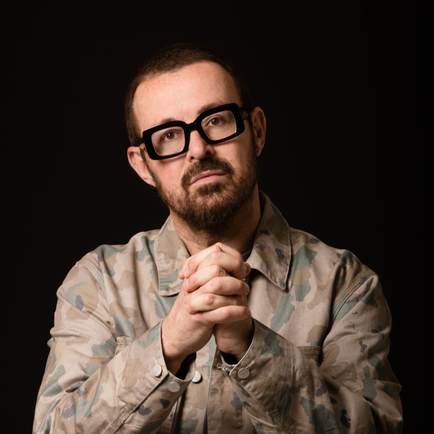 A photo of DJ Judge Jules. He is wearing a camouflage jacket with his hands clasped together. He has chunky black glasses on. The photo is taken against a plain black background. 